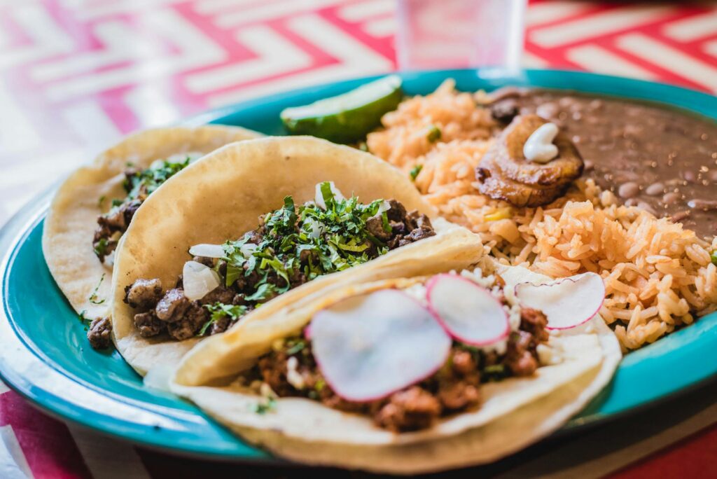 A vibrant turquoise plate holding three authentic street tacos topped with cilantro and radish, served alongside Mexican rice and refried beans for a Cinco de Mayo feast.