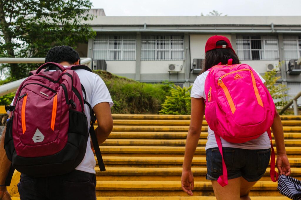 Two students seen from behind, one with a maroon backpack and one with a bright pink backpack, walking up yellow stairs towards a school building.