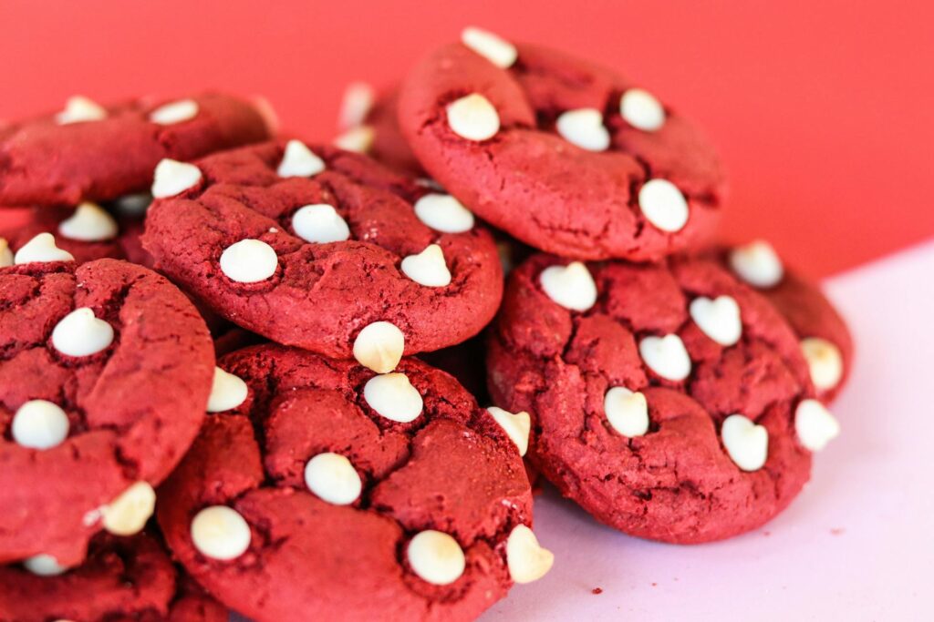 A close-up pile of homemade red velvet cookies studded with white chocolate chips, sitting on a pink and red background
