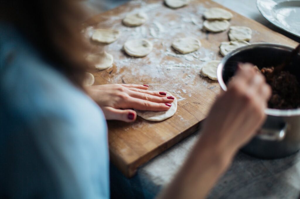 Close-up of hands with red nail polish preparing empanadas: placing filling on dough circles on a wooden cutting board

