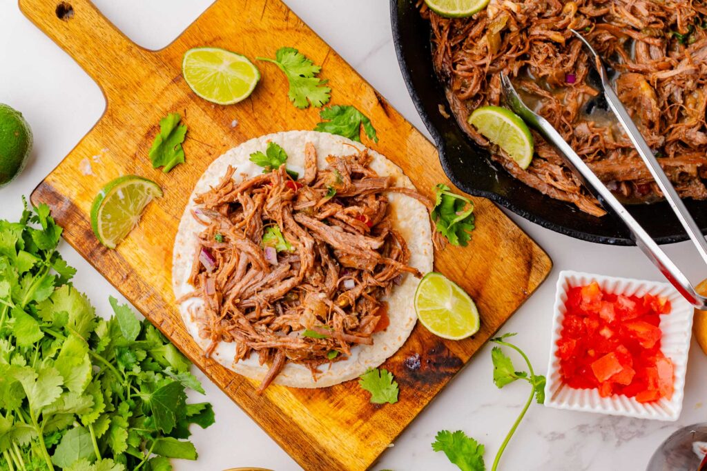 Delicious pulled pork taco on a fresh tortilla, served on a wooden cutting board with lime wedges, fresh cilantro, and diced tomatoes. In the background, a cast iron skillet filled with tender shredded pork, ready for more tacos
