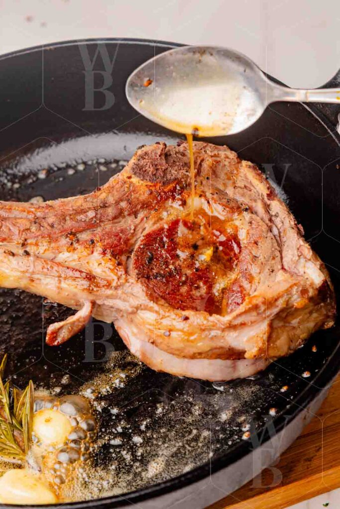 Process shot of a tomahawk steak being basted with a spoon in a cast iron pan